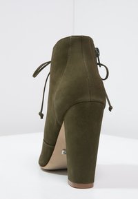 Botín de ante verde de tacón alto con punta redonda, detalle de cordones en la parte trasera, textura suave y tacón grueso.