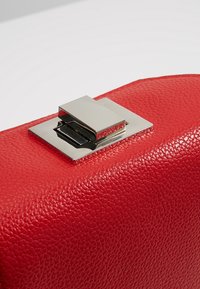 Sac à main en cuir rouge avec une finition texturée, doté d'un fermoir carré métallique et d'un accent lisse et poli sur le fermoir.