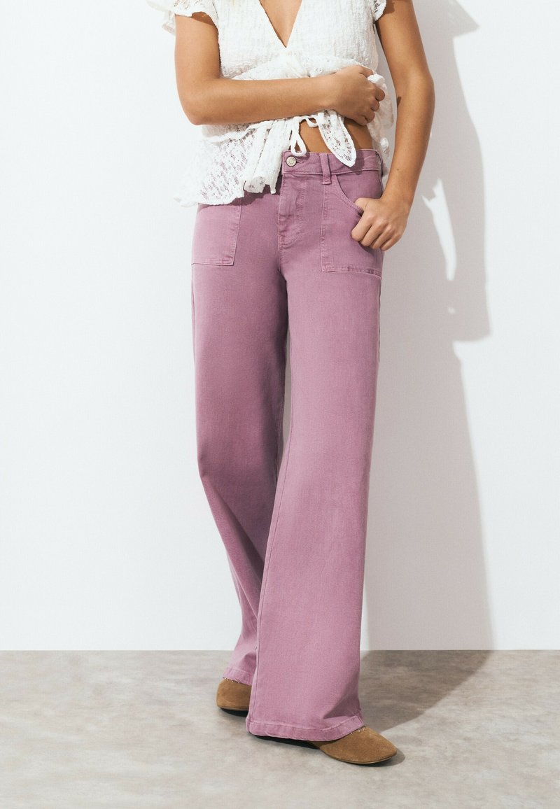 Mujer con pantalones rosas de tiro alto y pernera ancha, blusa corta de manga corta de encaje blanca atada, y zapatos marrones tipo slip-on, de pie contra una pared blanca.