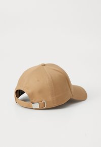 Tan baseball cap featuring a rounded crown, stitched accents, and a silver adjustable strap at the back. Smooth fabric texture.