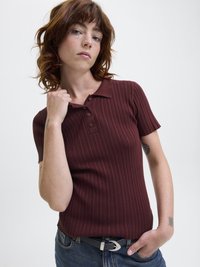 Ung person med skulderlangt brunt hår iført ribbet kortermet maroon polo-skjorte og blå jeans, poserer med den ene hånden på kragen og den andre i lommen.
