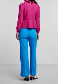 Top peplum en maille fuchsia avec un col à volants et une ouverture en forme de clé à l'arrière, associé à un pantalon bleu ajusté avec des fentes sur les côtés et des sandales à talons carrés noires.