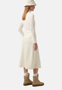 Cremfarbene, gesteppte Langarmkleid mit langen Ärmeln, ausgestattet mit einem Taillengürtel und seitlichen Taschen, kombiniert mit beigen Knöchelstiefeln und einem passenden Hut.