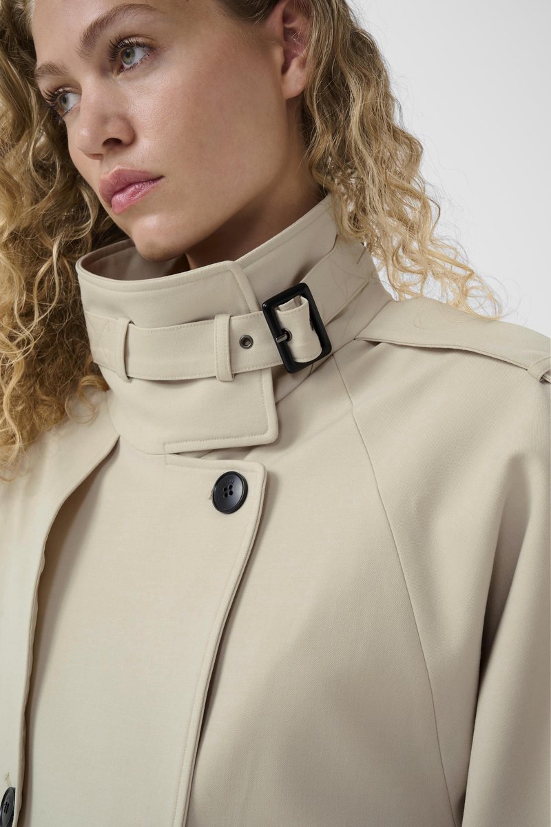 Femme aux cheveux blonds bouclés portant un trench-coat beige à col montant avec un détail de boucle noire, regardant pensivement sur le côté.