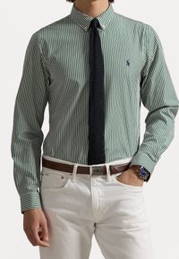 Grün-weiß gestreiftes Hemd mit Knopfleistenkragen, kombiniert mit einer marineblauen, strukturierten Krawatte und weißen Hosen, akzentuiert durch einen braunen Gürtel.