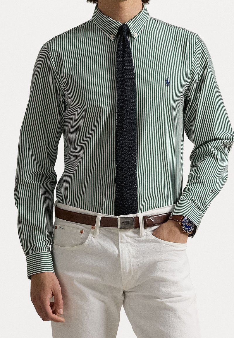 Grün-weiß gestreiftes Hemd mit Knopfleistenkragen, kombiniert mit einer marineblauen, strukturierten Krawatte und weißen Hosen, akzentuiert durch einen braunen Gürtel.