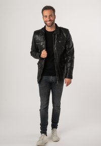 Giubbotto di pelle nera con numerose tasche e bottoni, abbinato a una t-shirt nera, jeans scuri e scarpe da ginnastica bianche. Sfondo neutro.
