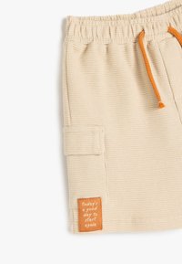 Beige shorts med vaffeltekstur, elastisk talje, orange snor og et patch med tekst, en sidelomme og opfoldet kant.