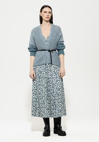 Himmelblau, gestrickte Strickjacke mit V-Ausschnitt und aufgerollten Ärmeln, tailliert, über einem floral gemusterten, bodenlangen Kleid mit Gänseblümchen.