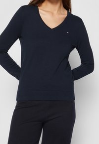 Pull marine à manches longues avec col en V, fabriqué en tissu tricoté doux, présentant un petit logo de la marque sur la poitrine et un ourlet côtelé ajusté.