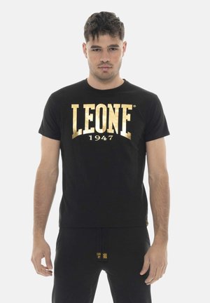 Jonge man met een zwart T-shirt met "LEONE 1947" in grote gouden letters, staand tegen een effen lichte achtergrond.