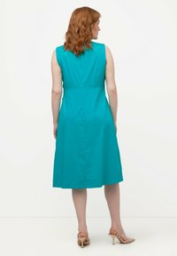 Un abito senza maniche di colore teal, lungo fino al ginocchio, con corpetto aderente e gonna svasata. Tessuto liscio e dettagli di cucitura discreti sul retro.
