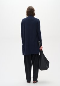 Marineblå langermet topp med krage, paret med løse svarte bukser. Personen holder en strukturert svart totebag. Bakgrunnen er hvit.