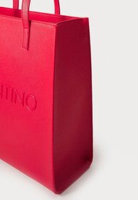 Röd syntetisk tote-väska med texturerad yta, med dubbelhandtag och präglad logotyp på framsidan. Fyrkantig form med platt botten.