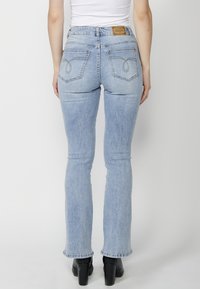 Pantalones de mezclilla acampanados de color azul claro con cintura alta, que cuentan con un parche de cuero marrón y bolsillos traseros cosidos, combinados con tacones negros.