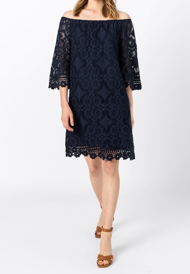 Femme en robe en dentelle bleu marine à épaules dénudées avec manches trois-quarts et sandales marron à bride autour de la cheville, marchant sur fond blanc.