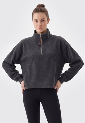 Kvinde iført en mørkegrå halvlukket pullover og sorte leggings, står med hænderne i siden mod en ensfarvet lys baggrund.