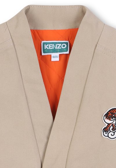 Veste beige avec un intérieur matelassé orange. Présente une étiquette Kenzo Kids et une broderie de tigre sur le côté gauche. Texture lisse.