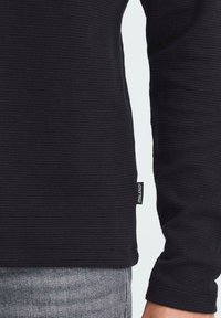 Črna strukturirana dolga majica z rebrom in majhno oznako blagovne znamke na strani, v kombinaciji s sivim džinsom.