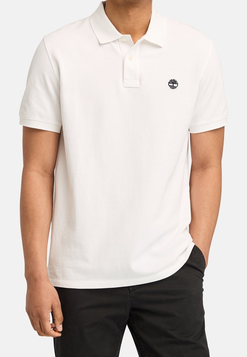 Homme portant un polo blanc à manches courtes avec un petit logo arbre noir sur la poitrine et un pantalon noir.