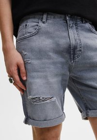 Pantalones cortos de mezclilla grises con dobladillo deshilachado, que presentan un ligero desgaste en la parte frontal. Con puños enrollados y diseño estándar de cinco bolsillos.