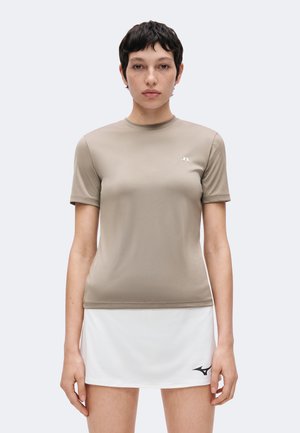 Femme aux cheveux courts et foncés portant une chemise beige à manches courtes et une jupe blanche avec des logos sportifs, debout devant un fond uni.