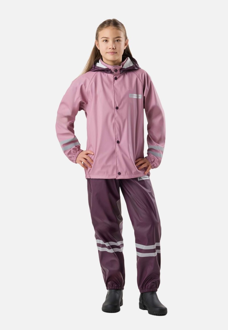 Enfant debout dans une veste et un pantalon imperméables roses et violets avec des bandes réfléchissantes, portant des bottes en caoutchouc noires, mains sur les hanches.