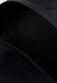 Juodos audinio vidus krepšio, turintis lygią tekstūrą ir mažą įspaustą etiketę. Užtrauktukas palei kraštą apibrėžia atidarymą.