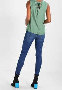 Zelená bezrukávová blůza s detailem uvázání na zadní straně, kombinovaná s tmavě modrými skinny džíny a černými kotníkovými botami s modrými blokovými podpatky.