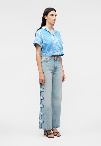 Γυναίκα που φορούν μπλε κοντομάνικο πουκάμισο με σχέδια, ανοιχτό μπλε τζιν παντελόνι με φαρδύ πόδι και μπλε διακοσμητικές πλευρικές ρίγες, και μαύρα σανδάλια.