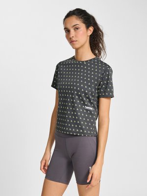 Vrouw met krullend haar naar achteren gebonden, draagt een zwart kort mouwig shirt met patroon en grijze aansluitende fietsbroek, staand tegen een witte achtergrond.