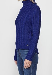 Pull en maille torsadée bleu royal avec col montant, présentant des motifs texturés et des fentes latérales, fabriqué en matière douce et épaisse.