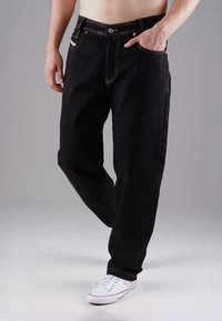 Černé džínové kalhoty s kontrastním oranžovým šitím, pěti kapsami a rovnými nohavicemi, v kombinaci s bílými teniskami.