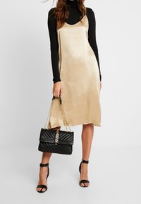 Žena v béžových šatech bez rukávů přes černý rolák, držící černou prošívanou kabelku a obutá v černých lodičkách na kotníkovém pásku.