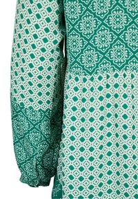 Blusa verde con motivo geometrico e maniche strutturate. Il tessuto ha una finitura liscia con disegni bianchi distintivi.