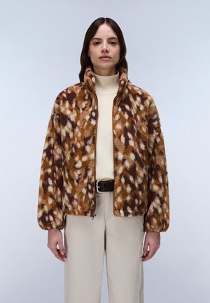 Kvinde iført en brun og beige mønstret fleecejakke, cremefarvet rullekravesweater og lyse bukser med sort bælte.