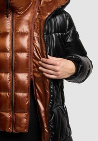 Zwarte pufferjack met een glanzende buitenkant, die een warme, bruine, gewatteerde voering blootlegt. Voorzien van dubbele ritsen en elastische manchetten.