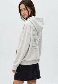 Giovane donna in una felpa grigia chiara con grafica di un cigno e testo sulla schiena, abbinata a una gonna nera, che guarda oltre la spalla.