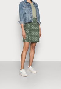 Džínová bunda přes zelenou skládanou sukni. Sukně má kruhový geometrický vzor. Modelka má na sobě bílé tenisky na jednobarevném pozadí.