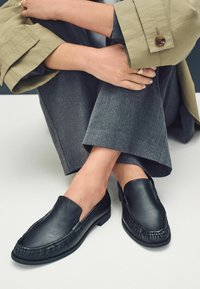 Mustad mustad nahast loafers, millel on sile viimistlus, silmapaistvad õmblused, ümar varvas ja madal blokknina. Kandmisel hallide pükstega.