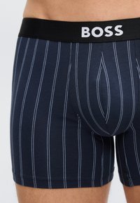 Κοντινό πλάνο ανδρικών μποξεράκ με ναυτικό μπλε χρώμα και λευκές κάθετες ρίγες, με μαύρη ζώνη μέσης που φέρει την ετικέτα "BOSS".