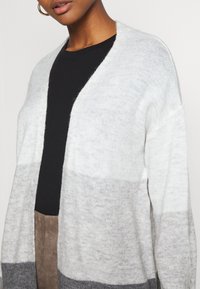 Cardigan grigio chiaro con una texture morbida. Presenta una vestibilità comoda, una apertura frontale e un design a gradiente che varia da grigio chiaro a grigio scuro.