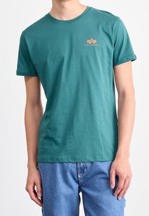 T-shirt à manches courtes couleur sarcelle en coton. Il présente un col rond et un petit logo orange sur le côté gauche de la poitrine. Coupe décontractée.