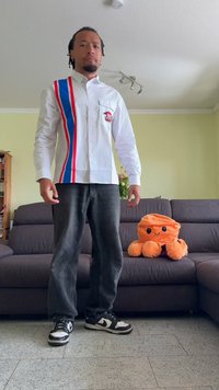 Camisa branca com riscas vermelhas e azuis, jeans pretos e ténis preto e branco. Apresenta um bolso frontal com um design impresso. Brinquedo de peluche laranja nas proximidades.