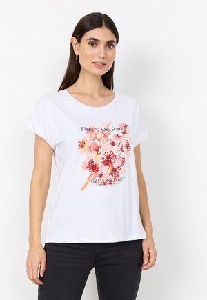 Donna che indossa una maglietta bianca con motivo floreale rosa e scritta "Fleurs De Paris" e pantaloni neri, in piedi davanti a uno sfondo bianco.