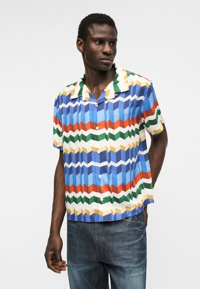 Άνδρας που φοράει πουκάμισο με κοντά μανίκια και πολύχρωμο γεωμετρικό ζιγκ-ζαγκ σχέδιο, συνδυασμένο με μπλε τζιν, στέκεται μπροστά σε απλό φόντο.