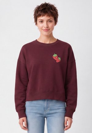 Junge Person mit kurzen Haaren, die einen burgunderfarbenen Pullover mit zwei kleinen Erdbeerabbildungen auf der Brust und hellblaue Jeans trägt.