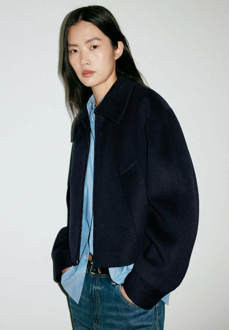 Jeune femme aux longs cheveux noirs porte une veste sombre oversize par-dessus une chemise bleue à rayures et un jean, debout les mains dans les poches.