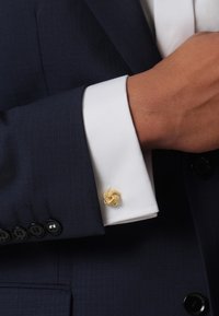 Navy suit with a white cuff featuring a gold knot cufflink. Textured fabric, contrasting colors, and distinctive cufflink design.