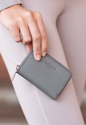 Grå læderpunge med lynlås, prydet med prægede logoer og sølvfarvet lynlåse, holdt i en hånd mod et lysfarvet leggings.